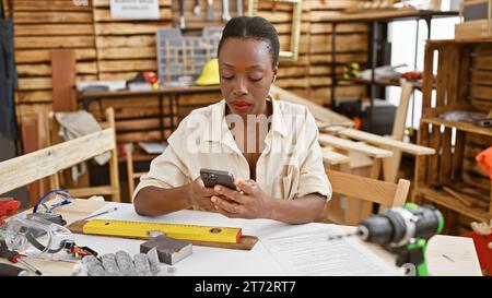 Serious african american female carpenter proficiently texting on smartphone while gracefully sitting at carpentry workshop table Stock Photo