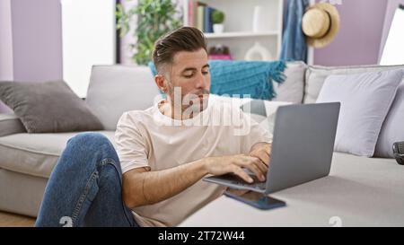 Attractive young hispanic man, concentrated and serious, sitting on the floor in his cozy apartment, working online on his laptop, deeply immersed in Stock Photo