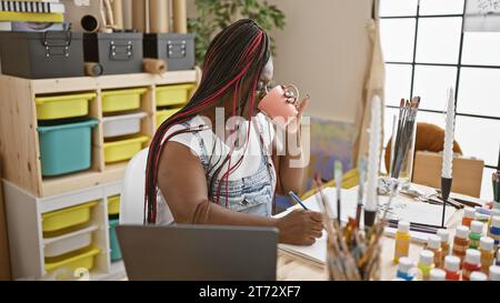 Beautiful african american woman artist painting a portrait, drawing on notebook whilst delightfully drinking morning coffee in her cozy art studio Stock Photo