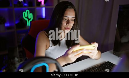 Gorgeous young hispanic woman streamer, passionately immersed in playing video game on smartphone in a dark gaming room, gamified ambiance at home Stock Photo