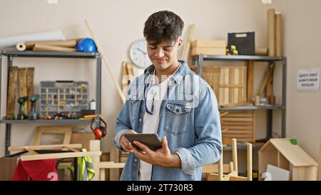 Smiling young hispanic man, a professional carpenter, engrossed in his carpentry work indoors, skillfully using a touchpad in his well-furnished works Stock Photo