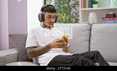 Handsome young latin man in a relaxed mood, sitting on his home sofa, intently listening to music through his smartphone's headphones, embodying a mod Stock Photo