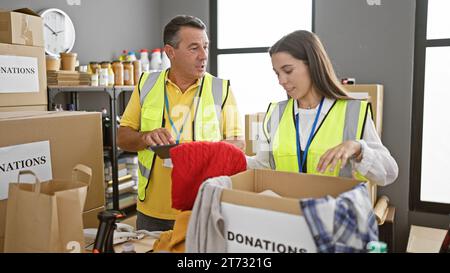 Smiling volunteers, man and woman, stand together in unity at the charity center - packaging clothes for a donation using their trusty touchpad. Stock Photo