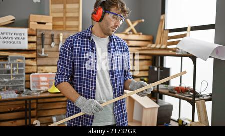 Handsome young adult, blond and bearded carpenter guy wearing safety headphones, professionally holding his wood plank in a vibrant carpentry studio i Stock Photo