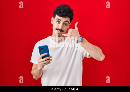 Hispanic man using smartphone over red background smiling doing phone gesture with hand and fingers like talking on the telephone. communicating conce Stock Photo