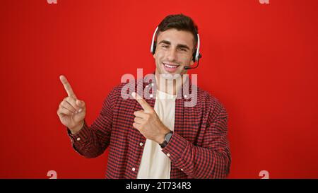 Enthusiastic young hispanic man wearing headset, working as a successful call center agent, cheerfully pointing to the side, presenting over a bold re Stock Photo