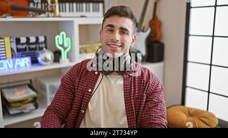 Confident young hispanic man smiling, wearing headphones at music studio, immersed in his music and song, embodying a happy performer and singer Stock Photo