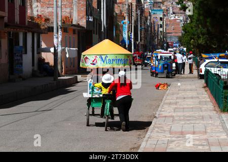 A woman and her daughter push a tricycle cart selling ceviche (raw, marinated fish, a famous typical Peruvian dish) along a street in Puno, Peru Stock Photo