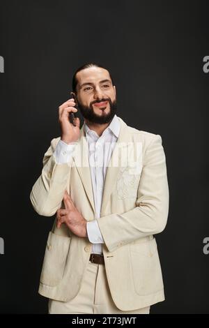 cheerful and bearded arabic man in white shirt and blazer talking on smartphone on black backdrop Stock Photo