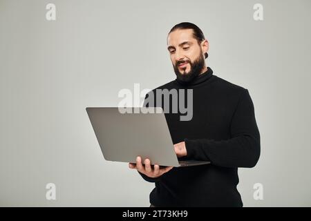 handsome and bearded arabic man in black turtleneck standing with laptop on grey background Stock Photo