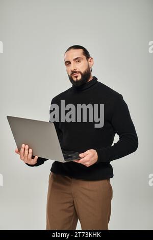 handsome and bearded arabic man in black turtleneck standing with laptop on grey background Stock Photo