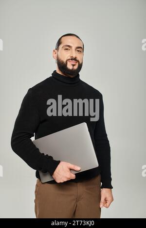 happy and bearded arabic man smiling and standing with laptop on grey background, freelancer Stock Photo