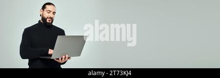 handsome and bearded arabic man in black turtleneck standing with laptop on grey background, banner Stock Photo