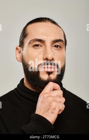 handsome middle eastern man in black turtleneck touching beard on grey background, confidence Stock Photo