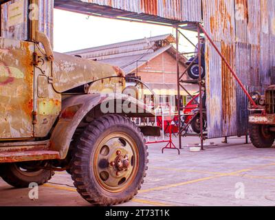 An antique military vehicle showing heavy rust parked in front of a weathered metal garage workshop in Southeast Asia. Stock Photo