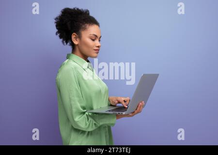 young pretty female model with curly black hair dressed in a green blouse and jeans works remotely using a laptop Stock Photo