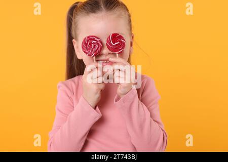 Happy little girl with bright lollipops covering eyes on orange background Stock Photo