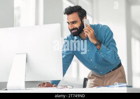 Smiling indian man talking on phone using computer Stock Photo
