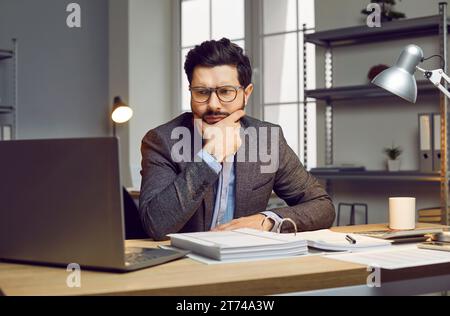 Portrait of serious, thoughtful, attractive, young businessman man with glasses, sitting at his workplace and looking at his laptop screen. Stock Photo