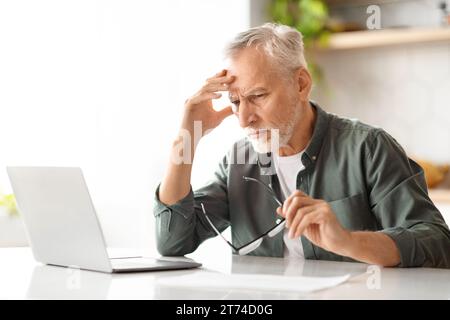 Stressed senior man using laptop while sitting at desk in kitchen Stock Photo