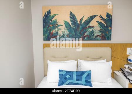 Interior view of hotel room featuring bed headboard adorned with decorative white and blue pillows and painting that hangs above bed on wall. Miami Be Stock Photo