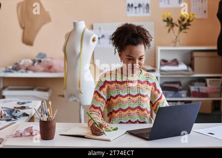 Young African American craftswoman watching online educational video and making notes in copybook while sitting by workplace in craft shop Stock Photo