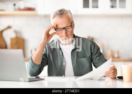 Home Office Paperwork. Senior Gentleman At Kitchen Reviewing Documents And Using Laptop Stock Photo