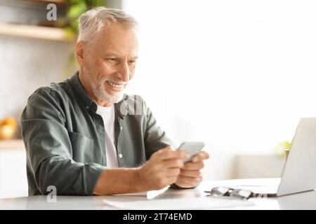 Happy Older Gentleman Using Smartphone While Sitting At Desk In Kitchen Stock Photo