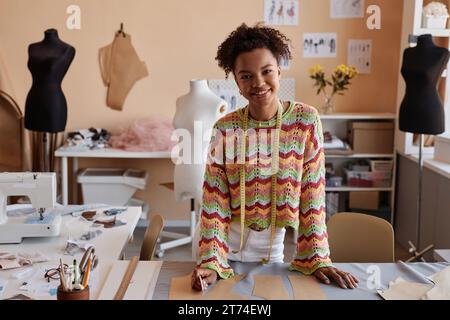 Young smiling craftswoman in casualwear standing by workplace in studio of fashion design or tailoring shop and looking at camera Stock Photo