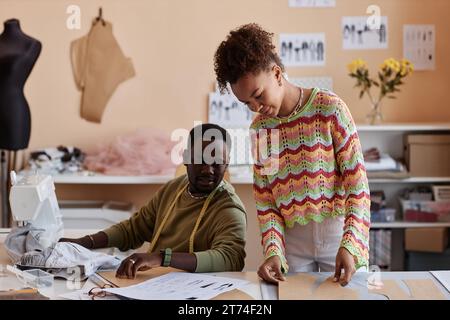 Two young African American tailors working over new fashion collection in studio or workshop while consulting about paper sewing patterns Stock Photo