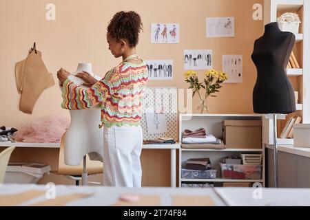 Young female stylist standing in front of mannequin in workshop and trying on new accessory or part of clothing item from new collection Stock Photo