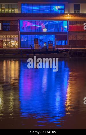 A tugboat moored up in front of an illuminated art work in M-Shed at night in Cumberland basin, Bristol docks, Bristol, Somerset, England, UK Stock Photo