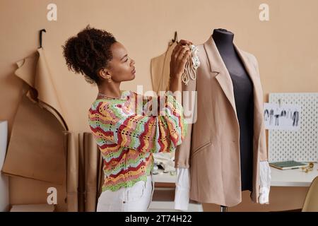 Side view of young female fashion designer sewing handmade decoration on beige blazer hanging on dummy while standing in workshop Stock Photo