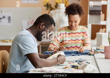 Side view of young creative male fashion designer making embroidery while sitting by workplace against female colleague in workshop Stock Photo