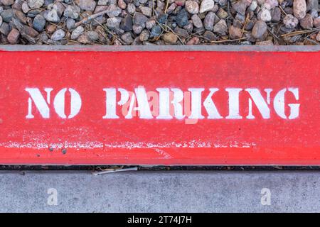 Close up of a red 'No Parking' sign with white lettering painted onto a sidewalk curb. Stock Photo