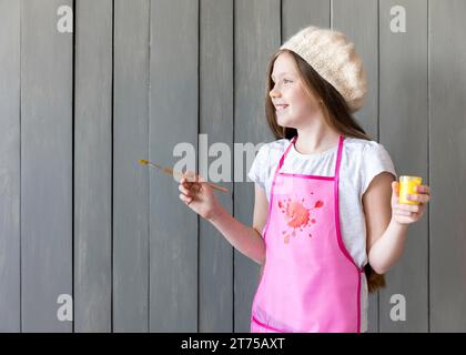 Side view smiling girl holding yellow paint bottle paintbrush hand standing against grey wooden wall Stock Photo