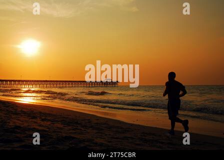 An adult man exercises and runs along the shore as the sun rises in the sky, near the pier in Myrtle Beach Stock Photo