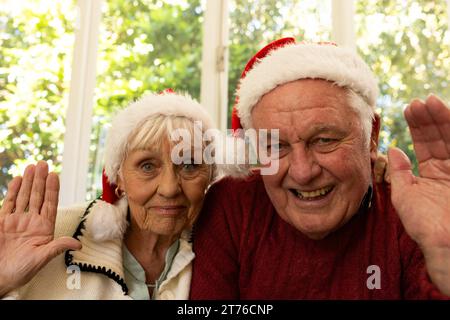 Happy caucasian senior couple in santa hats having christmas video call, waving in sunny room Stock Photo
