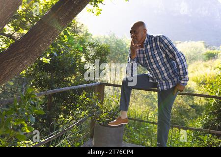 Pensive african american senior man sitting on fence in sunny garden, copy space Stock Photo