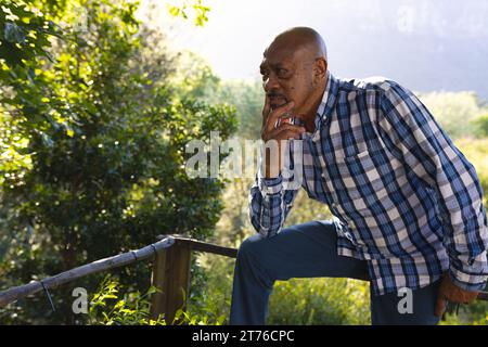 Pensive african american senior man sitting on fence thinking in sunny garden, copy space Stock Photo