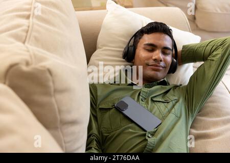 Happy biracial man lying on sofa using headphones at home Stock Photo