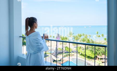 a Asian woman on vacation drinking coffee in the morning looking out the window over the beach and ocean during a luxury holiday Stock Photo