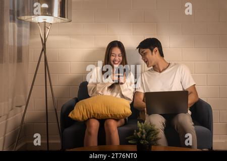 Young couple relaxing on couch with devices, woman laughs at phone content and sharing a funny moment together, cozy home setting Stock Photo