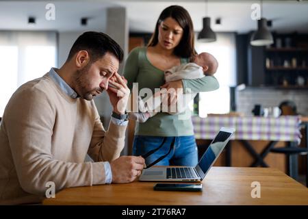 Stressful man trying to work on laptop with wife and children on background at home office. Stock Photo