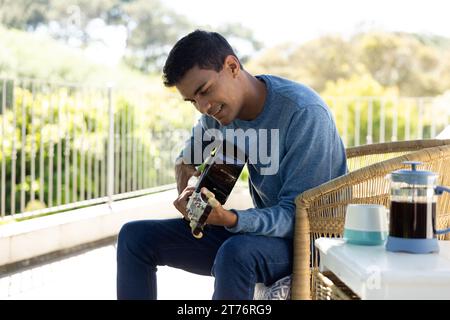 Happy biracial man sitting on balcony playing guitar at home Stock Photo