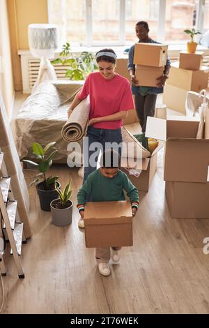 Cute little boy carrying packed box with household supplies while moving in front of his parents with rolled carpet and stack of packages Stock Photo