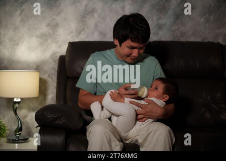 happy father feeding milk bottle to infant baby on a sofa in the living room at night Stock Photo