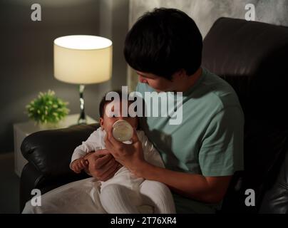 father feeding milk bottle to infant baby on a sofa in the living room at night Stock Photo