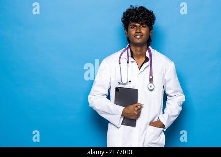 Revolution in healthcare, technologies in medical field. Cheerful positive handsome curly young indian doctor using digital tablet, isolated on blue b Stock Photo