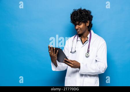 Revolution in healthcare, technologies in medical field. Cheerful positive handsome curly young indian doctor using digital tablet, isolated on blue b Stock Photo